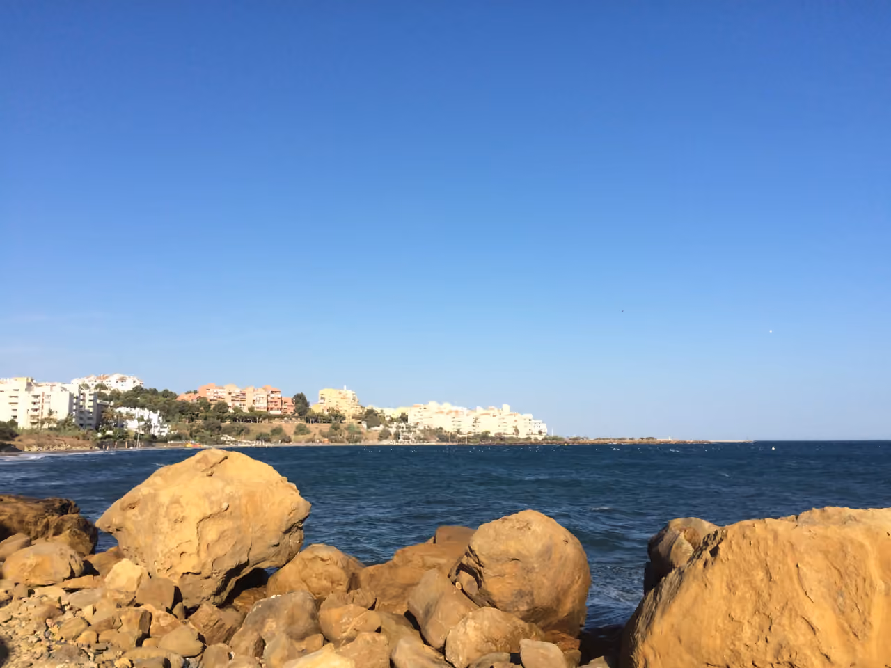 Estepona coastline near BAYSIDE HOMES, viewed from a rocky Mediterranean shore with residential buildings and the Mediterranean Sea in the background